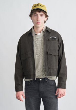 Mørke olivengrøn kort jakke med to forlommer, grå strikket sweater og mørke jeans. Gul kasket med logo foran.