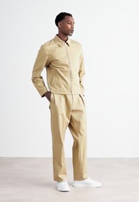Chaqueta con cremallera en tono beige y pantalones a juego hechos de un material suave; el modelo lleva zapatillas blancas; fondo liso con tonos neutros.