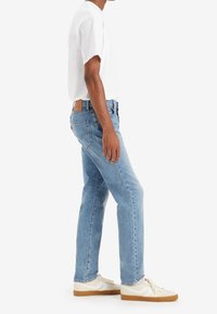 Jeans de mezclilla azul claro con corte recto, cinco bolsillos y un parche de cuero marrón en la parte de atrás. Llevados con una camiseta blanca simple y zapatillas.