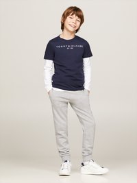 Dunkelblaues T-Shirt mit kurzen Ärmeln und weißem Logo, darüber ein weißes Langarmshirt, kombiniert mit hellgrauen Jogginghosen und weißen Sneakern.