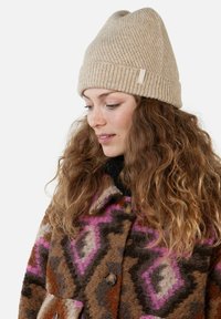 Beige knit beanie with ribbed texture, worn with a multicolored patterned jacket featuring brown, pink, and cream geometric designs.