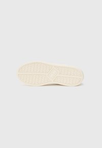 Hvit sneakersåle med teksturert gripe mønster, buet form og hevet logo detalj i midten. Glatt beige gummimateriale.