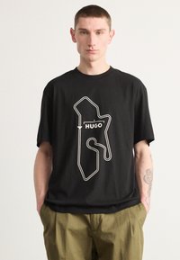 Melna kokvilnas t-kreklu ar baltu kontūrētu grafiku un zīmola nosaukumu "HUGO", kas skaidri redzams centrā.