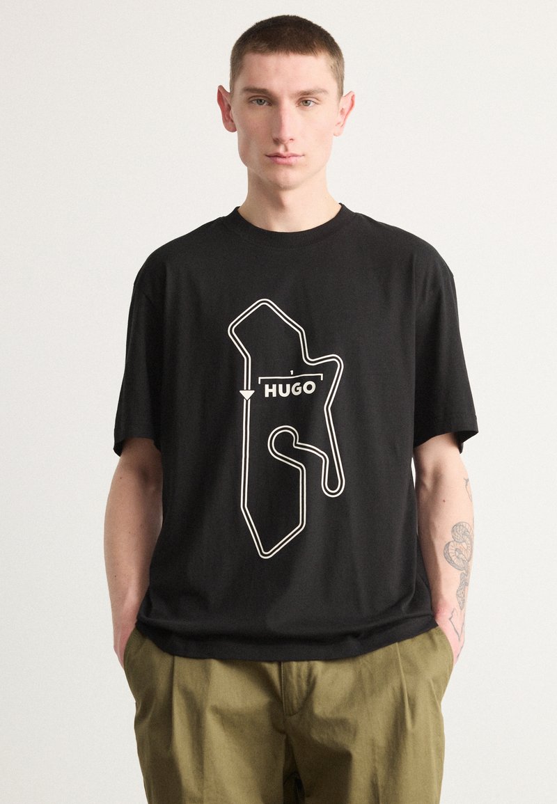 Melna kokvilnas t-kreklu ar baltu kontūrētu grafiku un zīmola nosaukumu "HUGO", kas skaidri redzams centrā.
