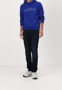 Blauer Hoodie mit weißem Logo, Fronttasche, kombiniert mit dunklen Jeans mit distressierten Details und grauen Sportschuhen.