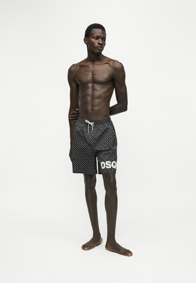 Homme debout pieds nus portant un short de bain noir à motifs avec un cordon blanc et des lettres blanches partielles sur la jambe droite sur fond blanc.