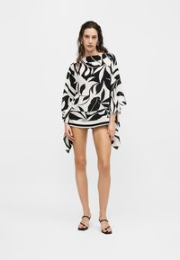 Νεαρή γυναίκα που φοράει μπλουζάκι σε μαύρο και άσπρο με υπερμεγέθη φυλλώδη μοτίβο και φαρδιά μανίκια, μαύρα σορτς και μαύρα σανδάλια με λουράκια, στέκεται απέναντι σε λευκό φόντο.