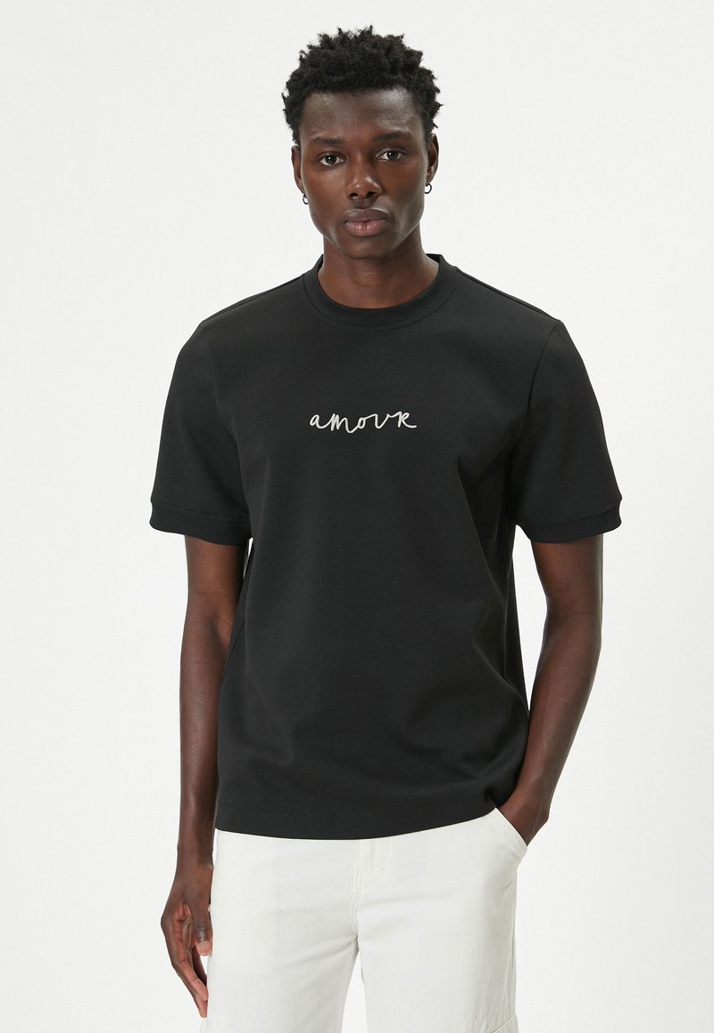 Sort bomulds t-shirt med korte ærmer, prydet med en hvid kursiv "amour" design på forsiden. Rund halsudskæring, afslappet pasform.