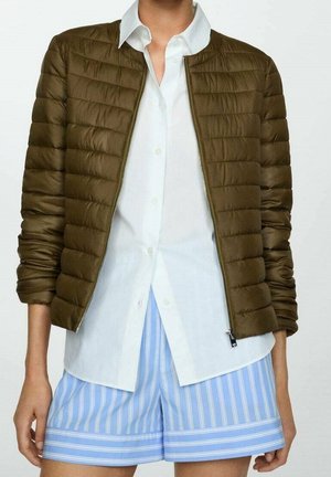 Persona con chaqueta acolchada con cremallera color oliva sobre una camisa blanca abotonada y pantalones cortos con rayas verticales azules y blancas.