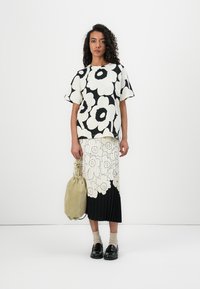 Bluse med blomstermønster i svart og hvitt, paret med et plisserte skjørt med store blomstermotiver, supplert med en lysegrønn veske og svarte sko.
