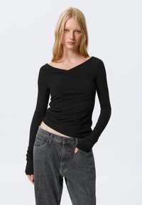 Sort langærmet top med crossover-udskæring, lavet af blødt, strækbart materiale. Kombineret med højtaljede, grå denimjeans.