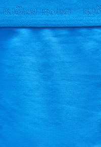 Κοντινή λήψη μπλε υφάσματος που δείχνει υφή πλέξης και ανεπαίσθητο ανάγλυφο κείμενο κατά μήκος του επάνω άκρου.