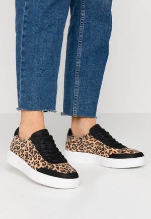 Persona con vaqueros azules recortados y zapatillas con estampado de leopardo con detalles negros y suelas blancas, de pie sobre un suelo claro.
