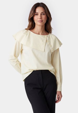 Woman with long brown hair wearing a cream blouse with a large squared collar and black pants, standing against a plain background.
