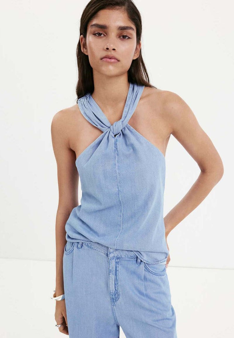 Jeune femme portant un débardeur en denim bleu clair noué au niveau du cou et un pantalon en denim taille haute assorti, posant la main sur la hanche.