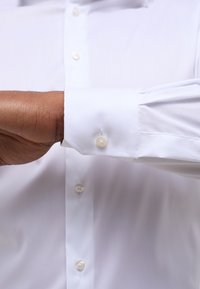 Camicetta bianca con una texture liscia e maniche lunghe. Presenta un polsino con bottone a un solo pulsante e una leggera lucentezza.