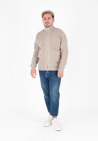 Beige zip-up vest met ribbeltextuur, gedragen over blauwe jeans en witte sneakers, staand voor een effen achtergrond.