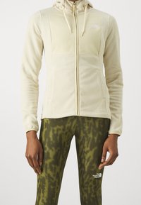 Chaqueta de forro polar beige claro con capucha, tela texturizada, cremallera delantera y logo en el pecho; combinada con leggings verdes con estampado.