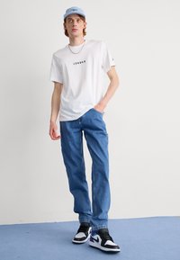 Camiseta de algodón blanca con texto negro, pantalones vaqueros de mezclilla azul y zapatillas negras con acentos morados. Una gorra azul claro completa el atuendo.