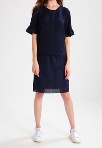 Modelka stoi w granatowym stroju, który składa się z plisowanej spódnicy i falbaniastej bluzki, w połączeniu z białymi trampkami, na jednolitym tle.