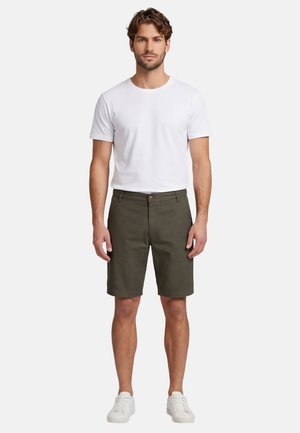 Man standing, wearing plain white short-sleeve t-shirt, olive green knee-length shorts, and white sneakers, facing forward on a white background.