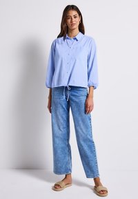 Camicia button-up azzurro chiaro con taschino sul petto e orlo con lacci, abbinata a pantaloni ampi in denim chiaro e sandali slip-on intrecciati color tan.