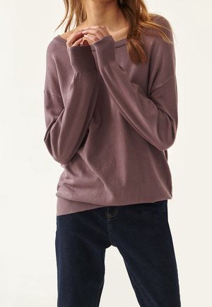 Femme portant un pull ample mauve à manches longues et un jean bleu foncé, les mains doucement jointes près de sa poitrine.