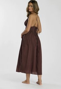 Bruine, mouwloze, middellange jurk met een strik op de rug, gerimpelde buste en een vloeiende rok van lichtgewicht stof.