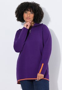 Een vrouw met krullend haar draagt een paarse trui met lange mouwen en oranje streepdetails, samen met een blauwe spijkerbroek, terwijl ze tegen een witte achtergrond staat.