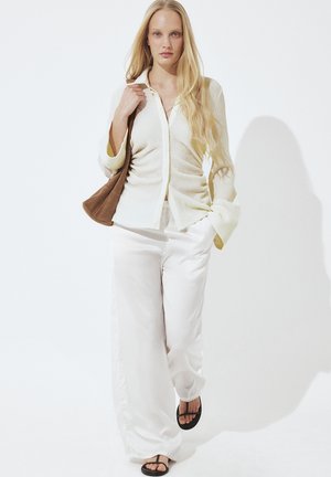 Žena s dlhými blond vlasmi, oblečená v krémovej textúrovanej košeli na gombíky, bielych nohaviciach s širokými nohavicami, čiernych sandáloch a sohnou hnedou kabelkou cez rameno.