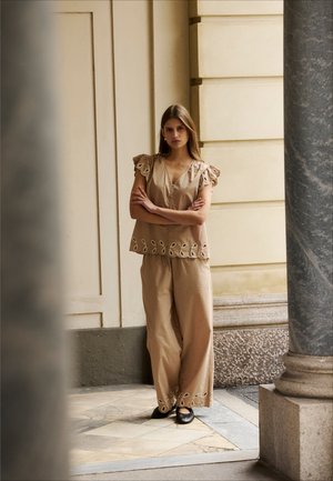 Kvinde stående med armene krydset iført beige outfit med hulmønster og sorte flade sko mod en sten- og cremefarvet væg.