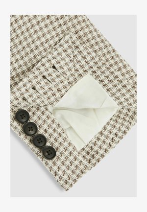 Struktureret stof i beige og brun zigzag mønster, med en hvid foring synlig i en lomme; fire sorte knapper på kanten.