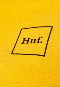 Gul tyg med en texturerad yta, med en svart fyrkantig kontur som innehåller ordet "Huf" tryckt med fet stil.