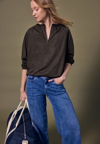 Brun zippered pullover med samlet halsudskæring, båret med blå vide jeans. Modellen holder en marineblå duffeltaske med cremefarvede stropper.