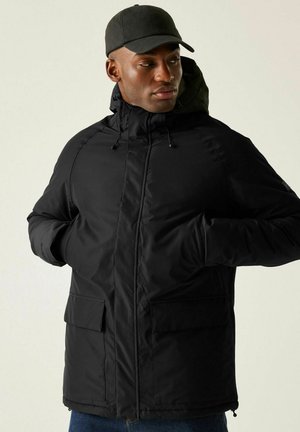 Man met zwarte winterjas met capuchon en zwarte pet, handen in de zakken, kijkt naar links tegen een lichte achtergrond.