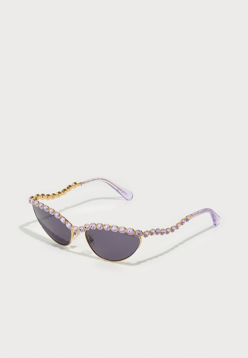 Occhiali da sole a forma di occhio di gatto con lenti scure, asticelle trasparenti in lavanda e una fila di strass viola lungo il bordo superiore e il tempio.