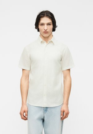 Νεαρός άνδρας με κοντά μαύρα μαλλιά, φορώντας ένα κοντομάνικο, κουμπωτό πουκάμισο εκρού και ανοιχτό μπλε τζιν, στέκεται μπροστά σε λευκό φόντο.