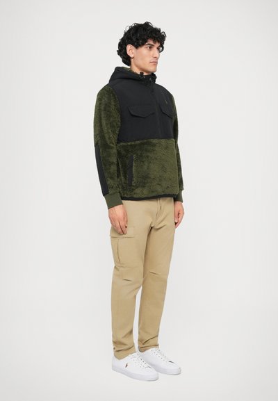 Casaco capuz verde e preto com textura felpuda, fecho de zíper e dois bolsos no peito, combinado com calças cargo bege e sapatilhas brancas.