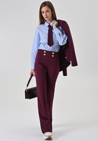 Pantalon tailleur bordeaux avec des boutons dorés, associé à une chemise bleu clair et une cravate. Tient un sac à main marron texturé. Baskets blanches.