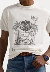 Kremowa bawełniana koszulka z nadrukiem w stylu vintage, przedstawiającym palmy, kotwice i chatki, ozdobiona ciemnoszarym tekstem oraz kwiatowym detalem.