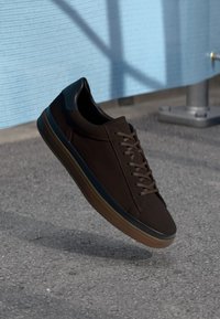 Bruna sneakers med en slät textilöverdel, rund tå och gummisula. Skosnörena är bruna, med en svart detalj på hälen.