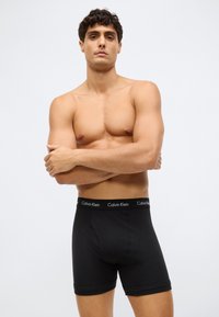 Homem musculoso com cabelo curto e escuro, usando boxers pretos da Calvin Klein, de pé com os braços cruzados em fundo branco simples.