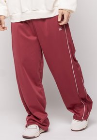 Person iført løse maroon bukser med hvid piping og hvide sneakers, der står foran en ensfarvet lys baggrund.