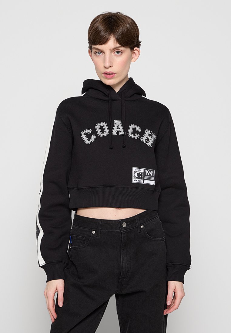 Schwarze, verkürzte Kapuzenjacke mit „COACH“ in strukturierten Buchstaben, seitlichen Streifen und einem Logo-Patch auf der Vorderseite. Der Stoff hat eine glatte Textur.
