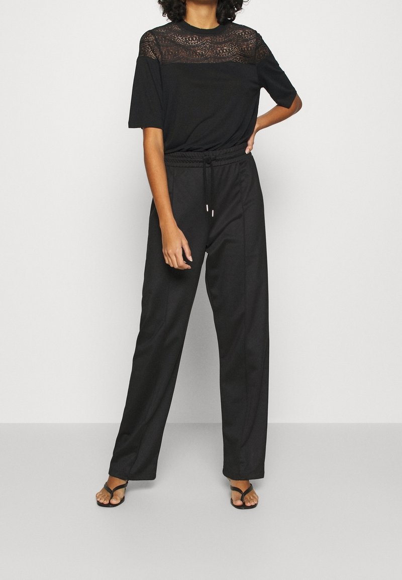 Femme portant un top noir à manches courtes avec empiècement en dentelle, un pantalon noir large à taille haute avec cordon de serrage, et des sandales noires à bout ouvert, debout avec la main sur la hanche.