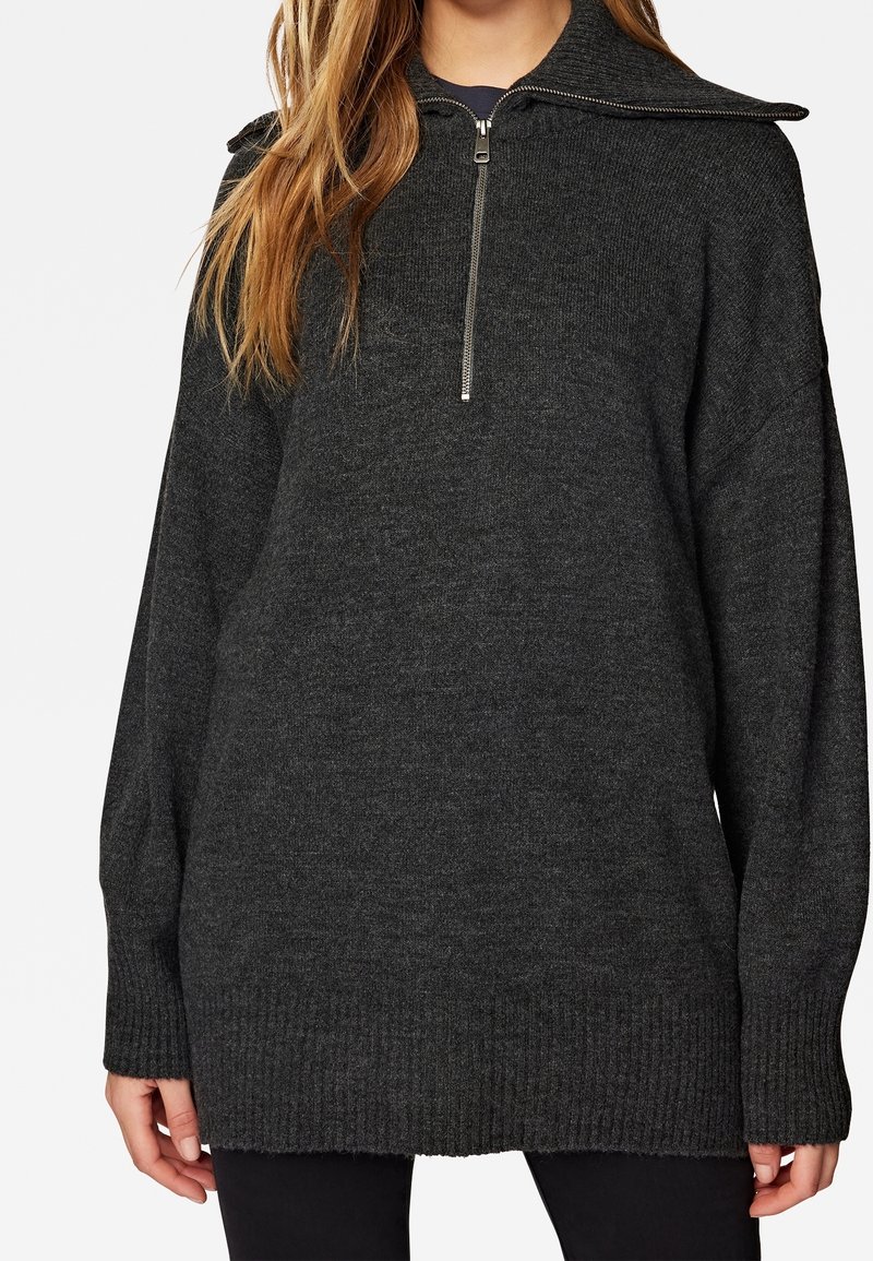 Pull en maille gris foncé à manches longues avec demi-fermeture éclair et col large, porté par une personne aux longs cheveux châtain clair et pantalon noir.