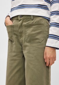 Παντελόνι ελιάς με φαρδιά μπατζάκια και μεγάλες πίσω τσέπες, κατασκευασμένο από ανθεκτικό ύφασμα. Συνδυάζεται με ριγέ πουκάμισο με μακριά μανίκια.