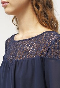 Blusa blu navy con pizzo finemente lavorato sulla spalla e tessuto trasparente e fluente sotto il petto.