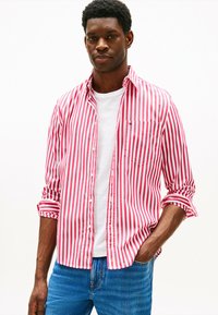 Camicia a righe rosse e bianche, maniche lunghe arrotolate fino ai gomiti, abbinata a una t-shirt bianca, indossata con jeans in denim blu.