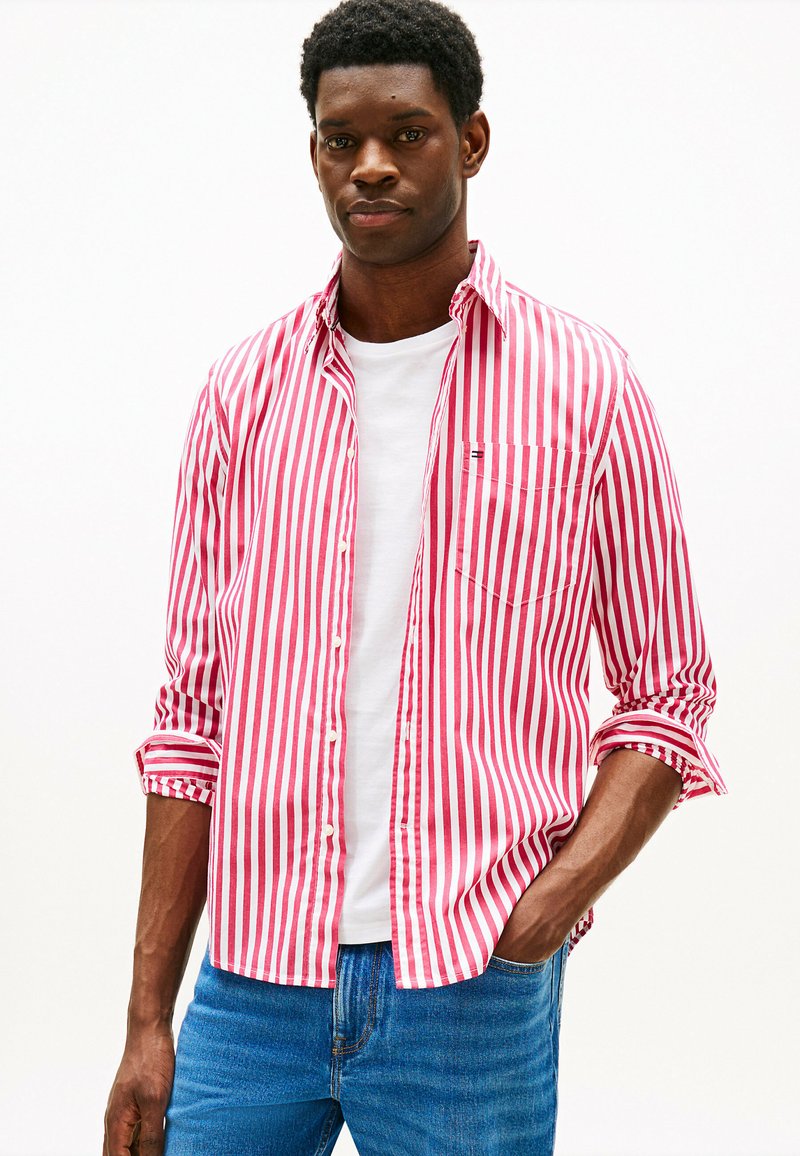 Camicia a righe rosse e bianche, maniche lunghe arrotolate fino ai gomiti, abbinata a una t-shirt bianca, indossata con jeans in denim blu.